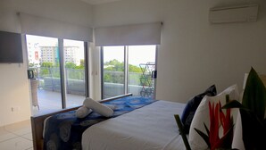 Un dormitorio con cama, balcón y vistas a edificios y vegetación, además de un aire acondicionado de pared.