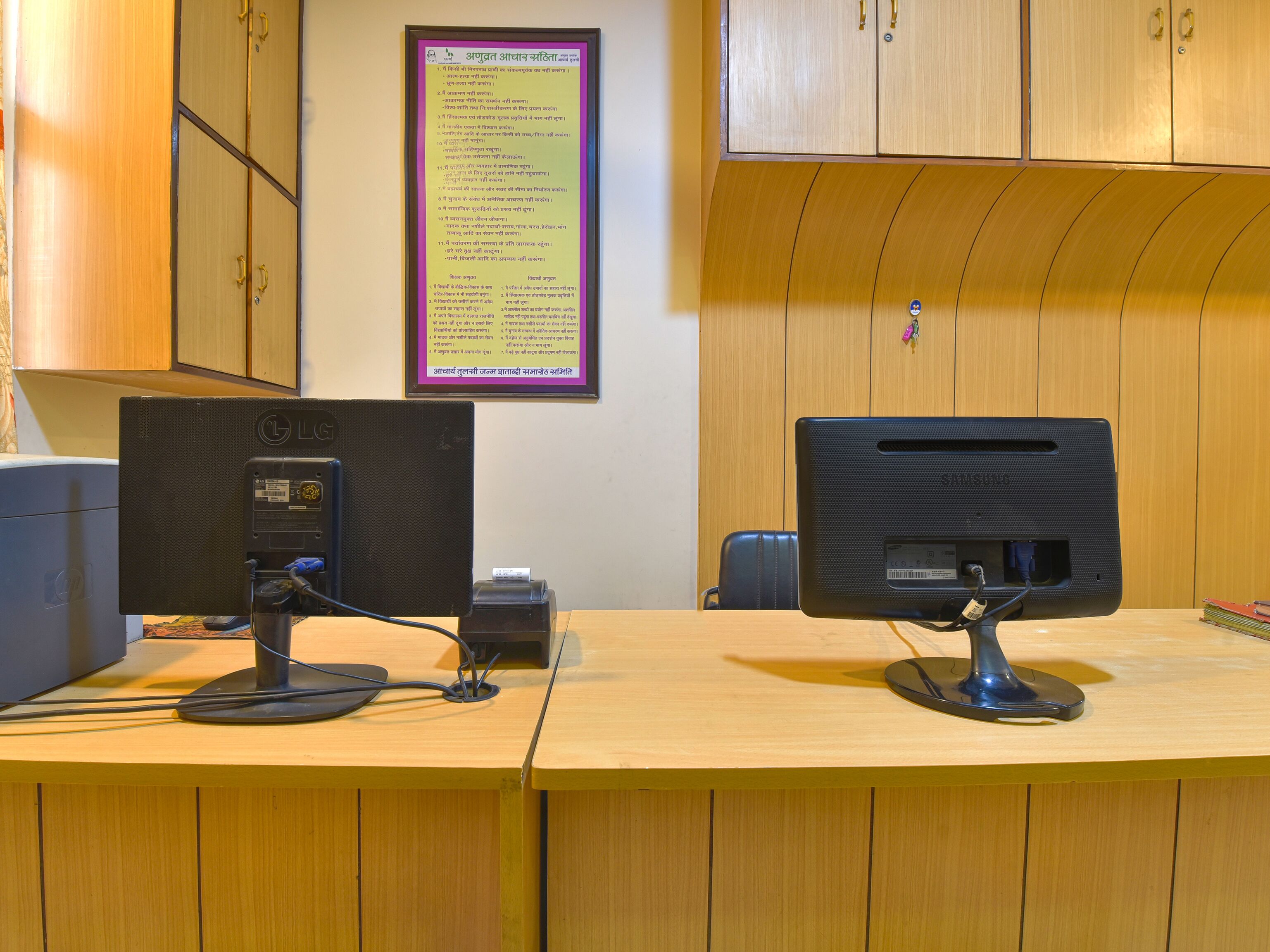 A computer desk with two monitors, a keyboard, and a mouse. There is a framed sign on the wall and wooden cabinets above the desk.