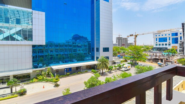 A modern building with a reflective glass facade, a spacious courtyard with palm trees, and a view of the surrounding urban area including construction cranes.