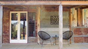 A covered outdoor area with two chairs and a table, a sliding glass door, and a corrugated metal wall.