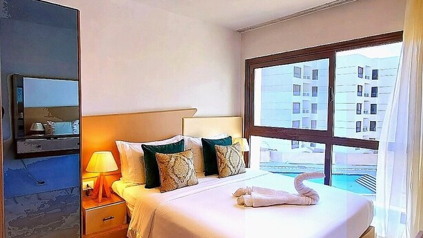 A hotel room with a bed, a nightstand, a lamp, a TV, a window with a view of buildings, and a ceiling light. - Your Home Away From Home at Porto Said Resort (Al-Manasrah)