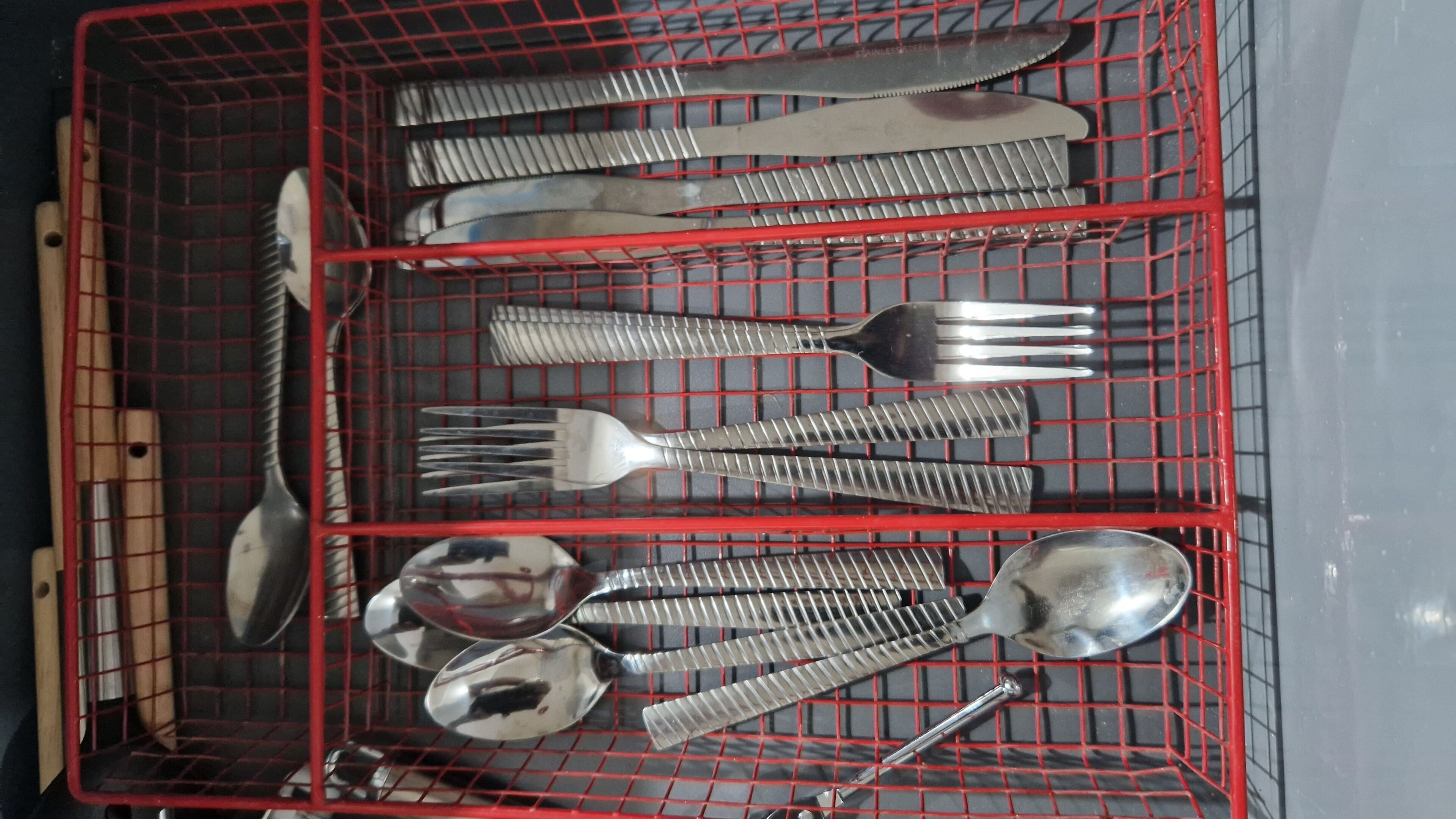 Een rode metalen mand met verschillende keukengerei, waaronder vorken, lepels en een mes.