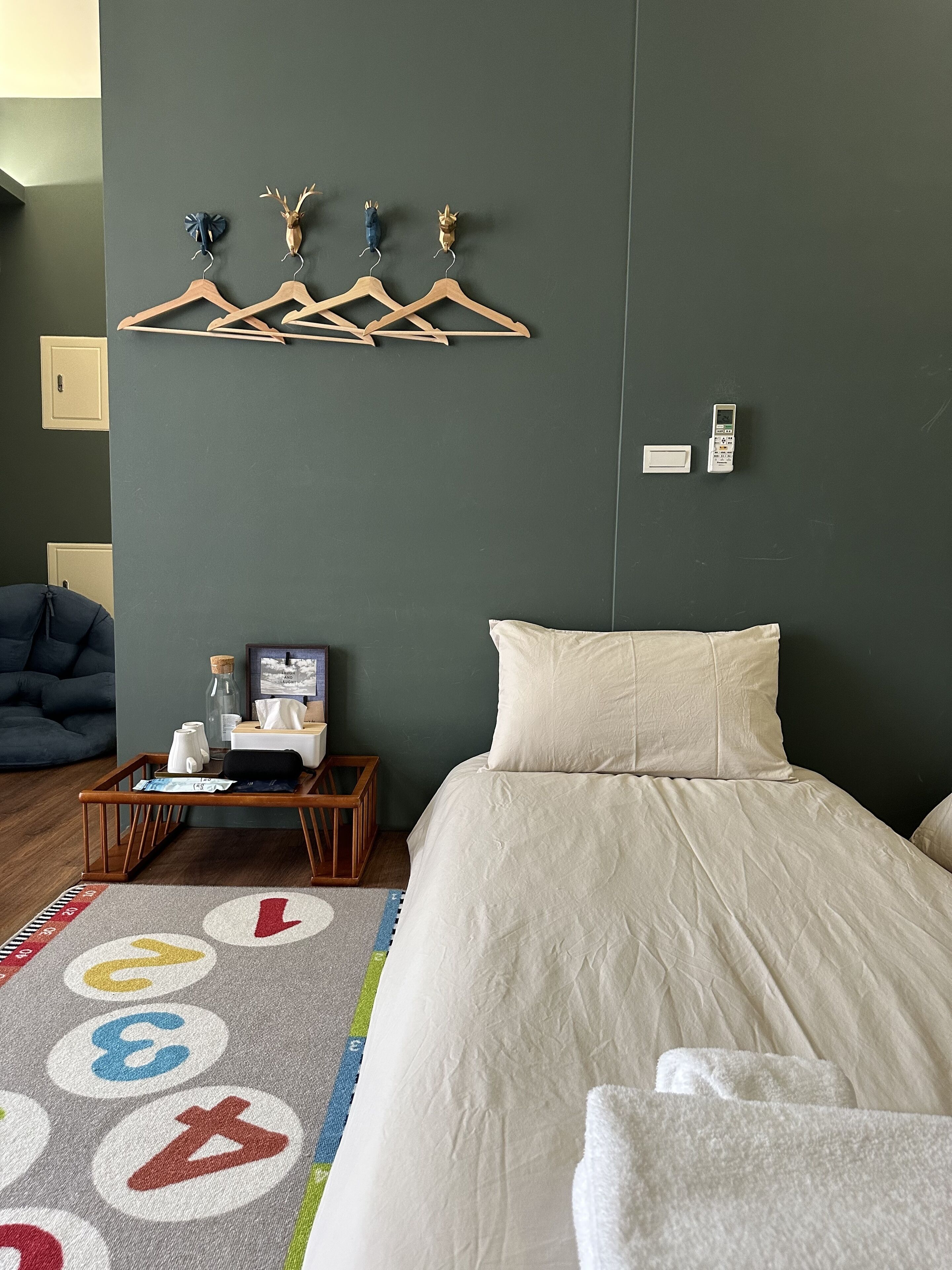 A bedroom with a bed, a bedside table with a lamp, a wall-mounted coat rack, and a small shelf with a phone and a box.