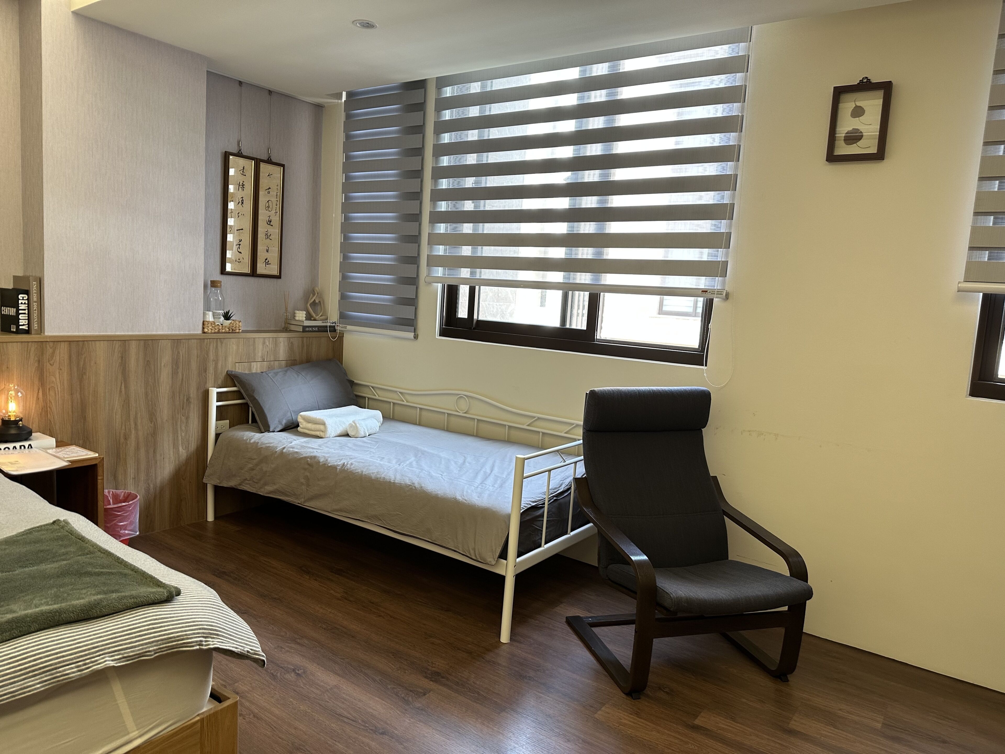 Un dormitorio pequeño y ordenado con una cama individual, una silla, una mesita de noche y una ventana con persianas.