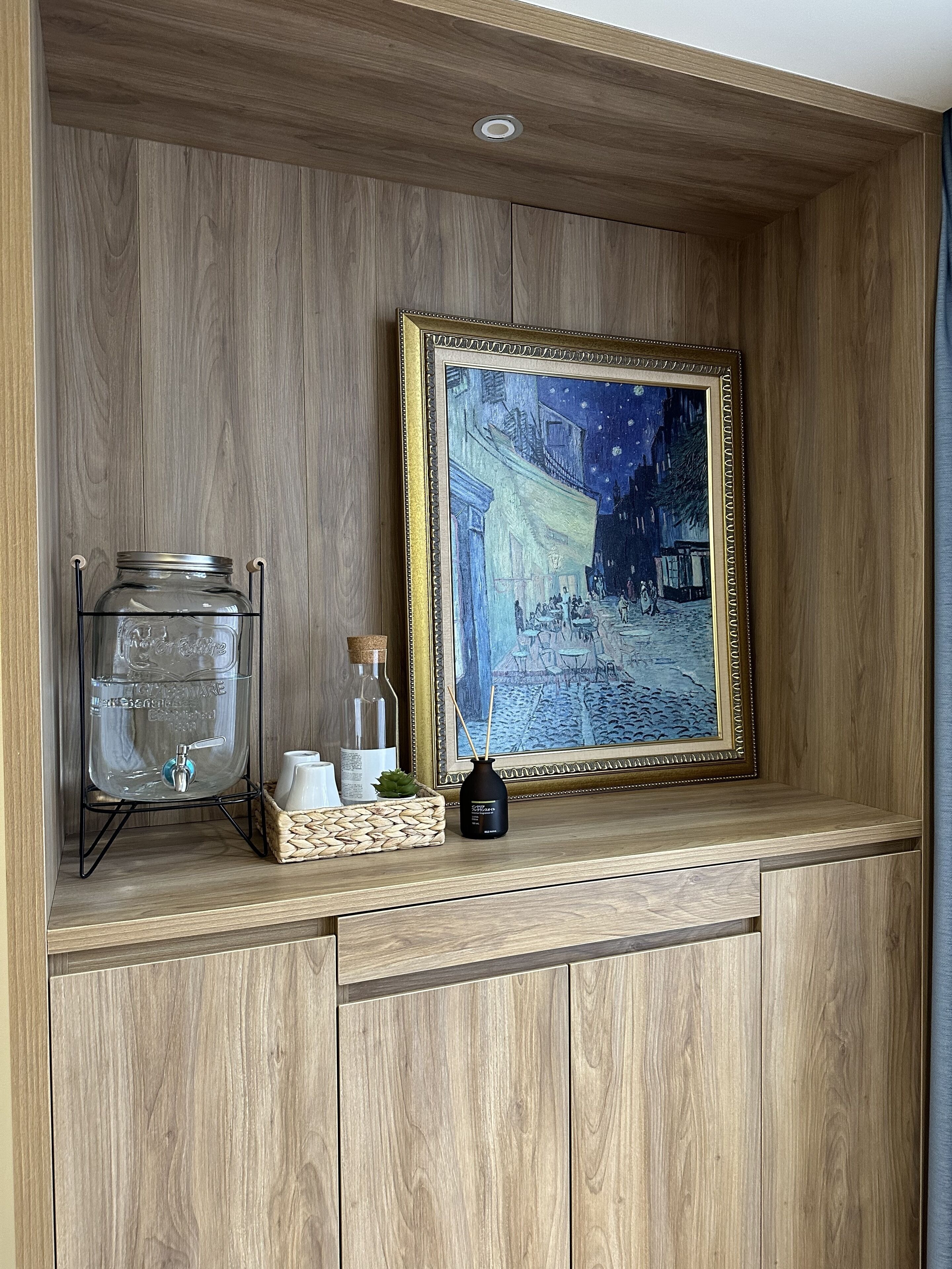 A wooden shelf with a water dispenser, a framed painting, and a reed diffuser.