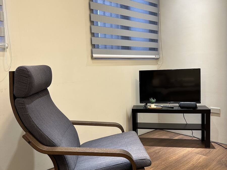 Ein modernes Wohnzimmer mit einem Liegesessel, einem Flachbildfernseher auf einem schwarzen Standfuß und einem Fenster mit Jalousien.