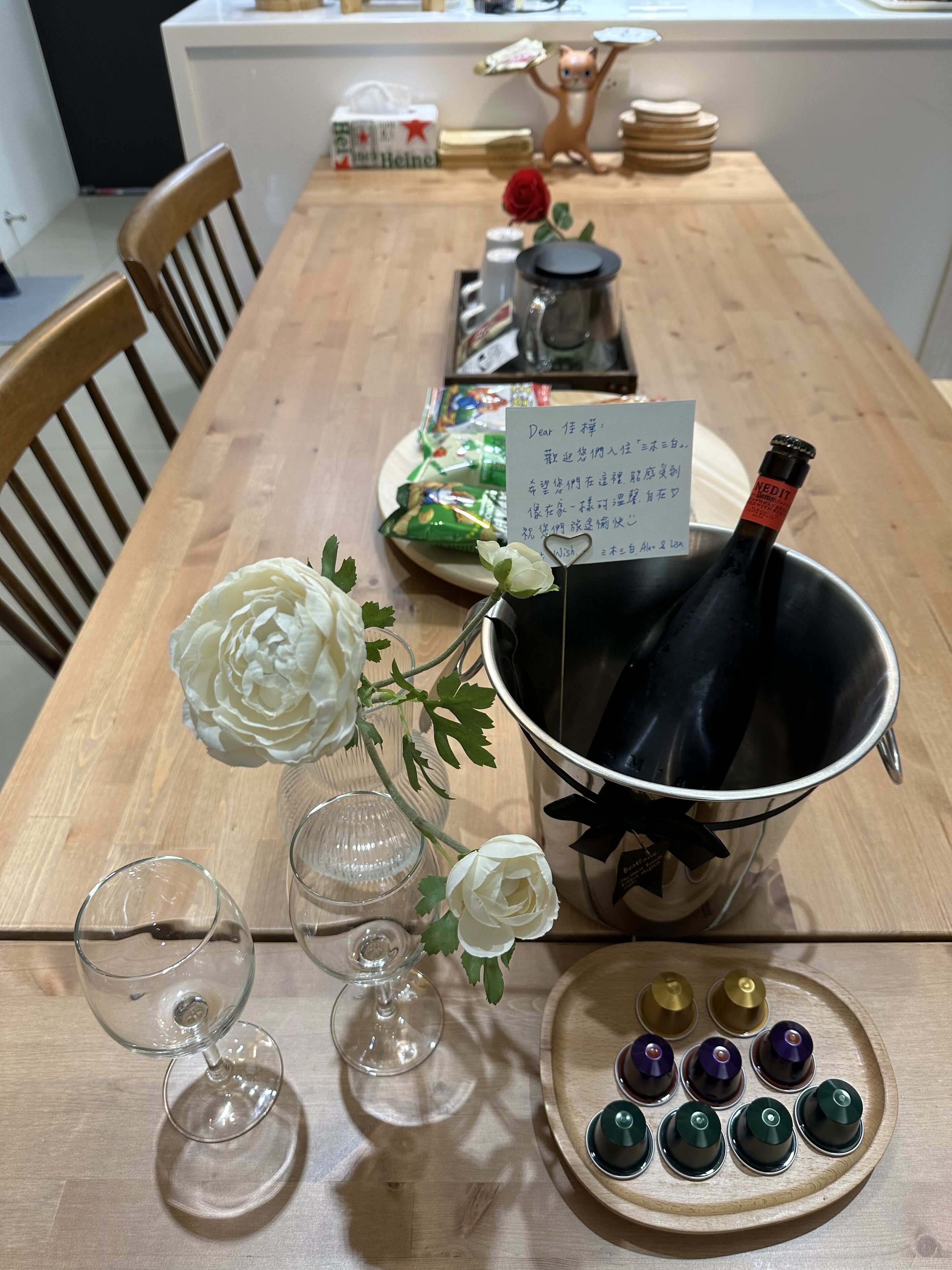 A wooden dining table with a bottle of wine, a flower vase, and a plate with small cups.