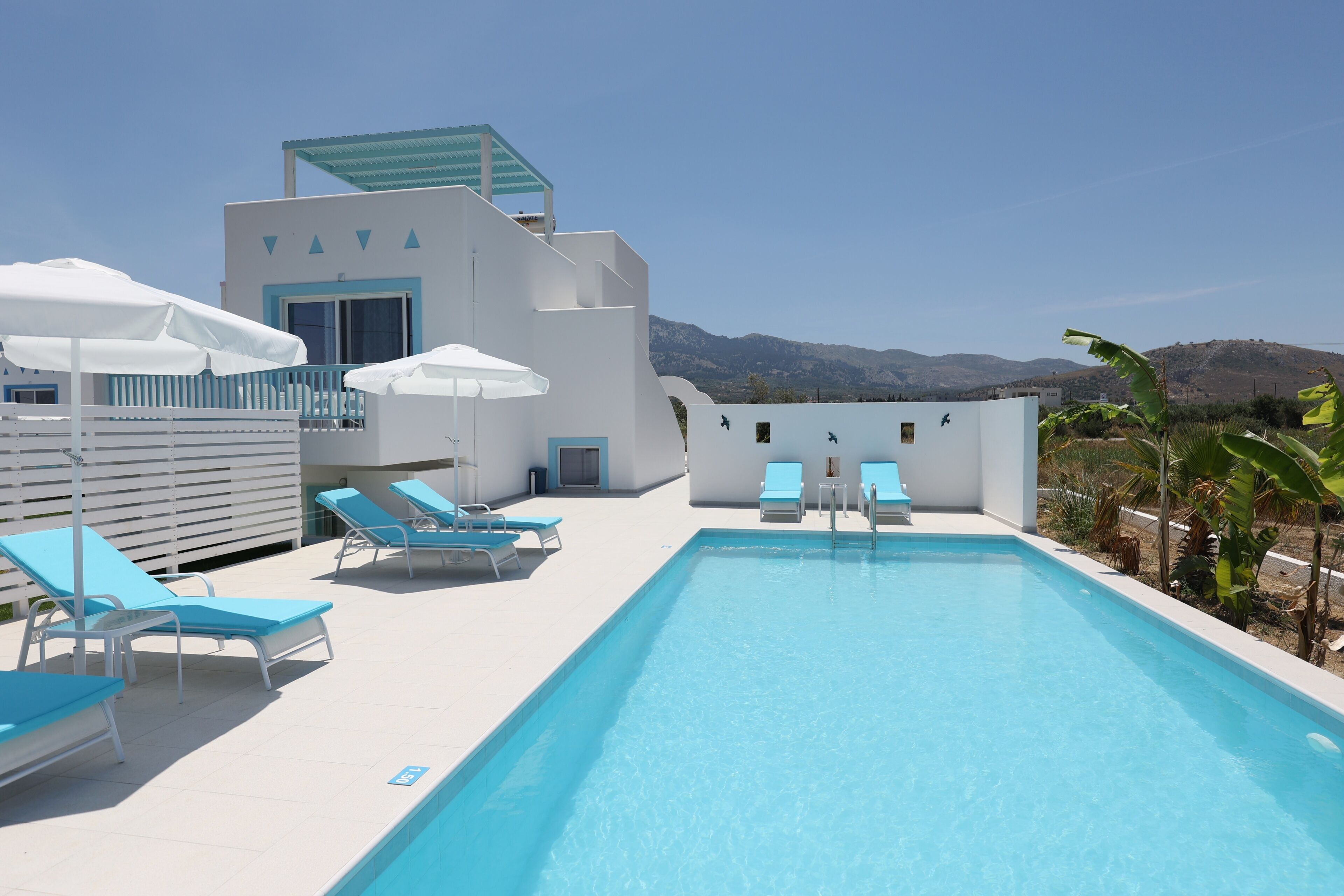 A pool area with blue lounge chairs, umbrellas, and a building with a white facade and blue accents.