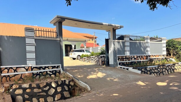 A gated entrance with a covered walkway, a parked car, and a building with a stone facade and metal railing. - Beautiful Family House (Kampala)