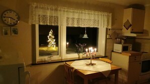Une cuisine chaleureuse avec une table à manger, une horloge, une fenêtre illuminée par un sapin de Noël et un four à micro-ondes.