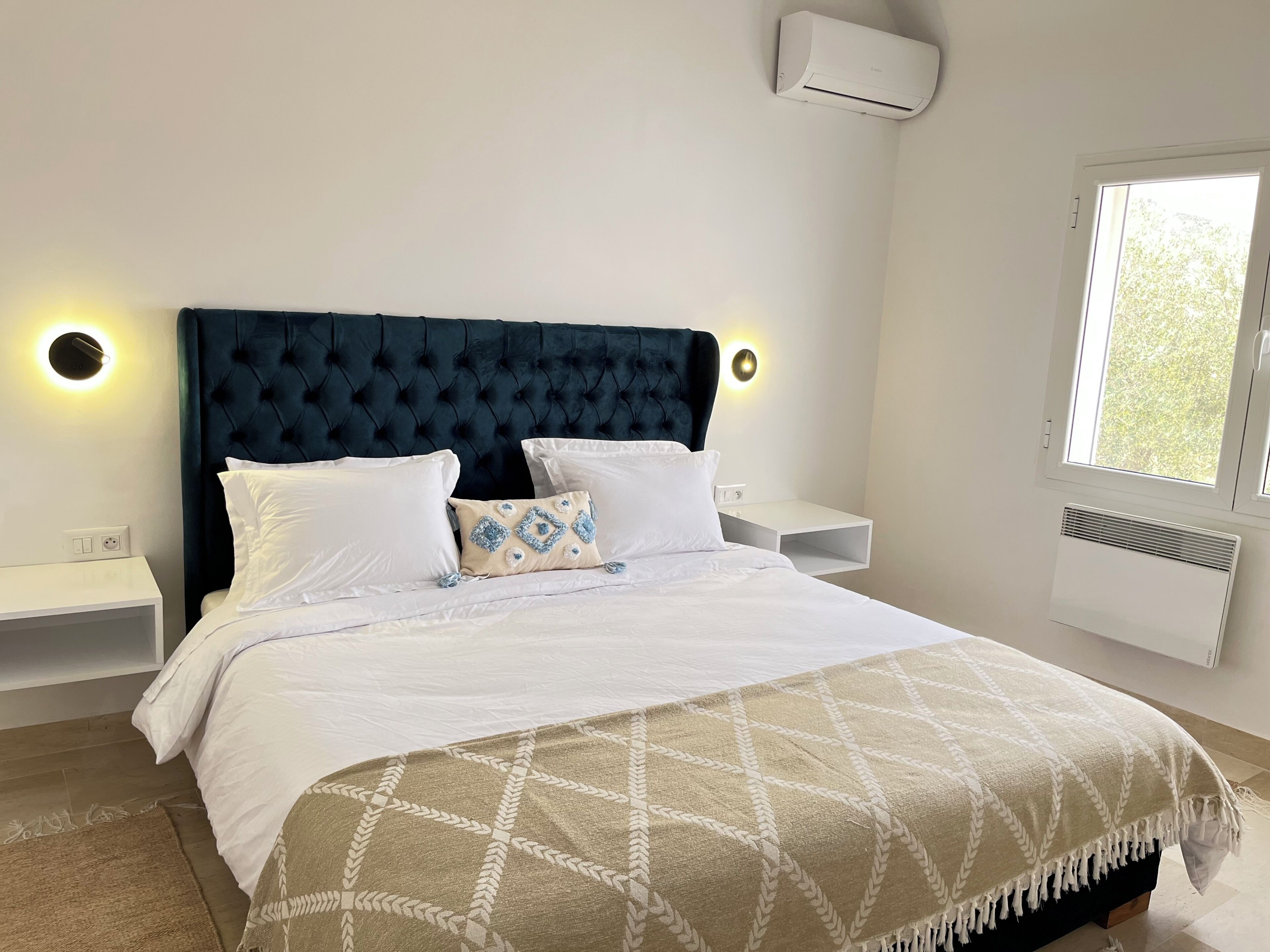A bedroom with a tufted headboard, white bedding, decorative pillows, bedside tables with wall-mounted lights, and a window with a view.
