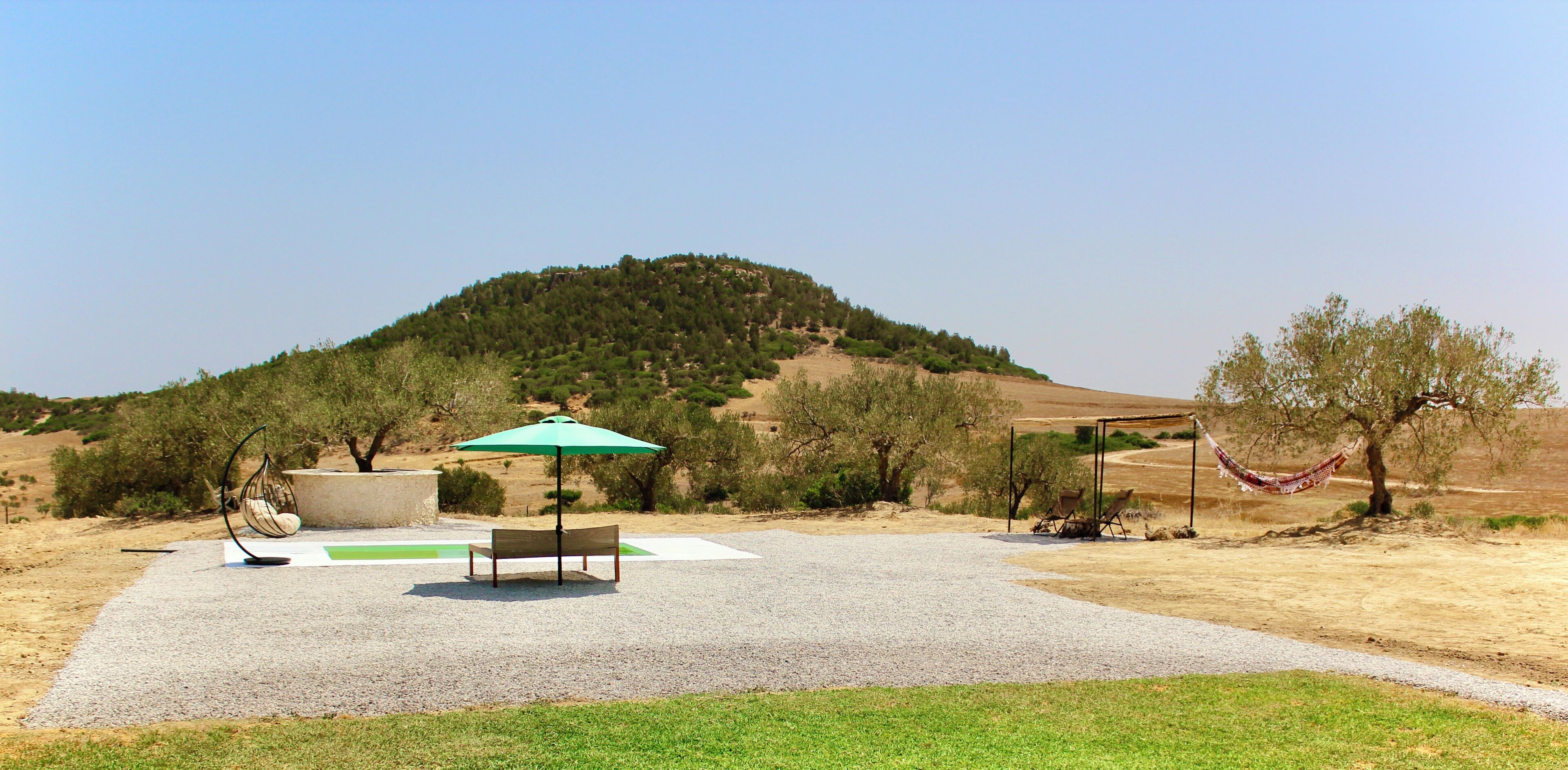A park with a bench, an umbrella, a hammock, and a hill in the background.