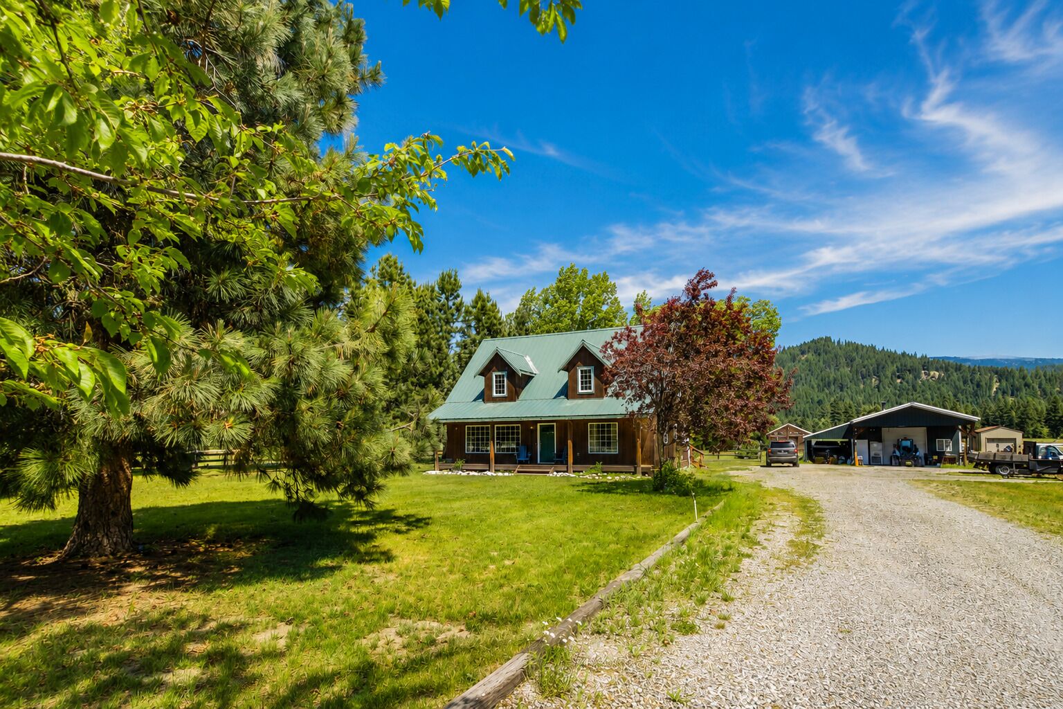 A lodge with a green roof, a large tree in the foreground, and a gravel driveway leading to the main building.