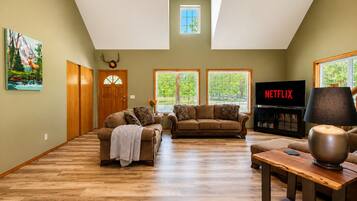 A spacious living room with a high ceiling, wooden flooring, and large windows.