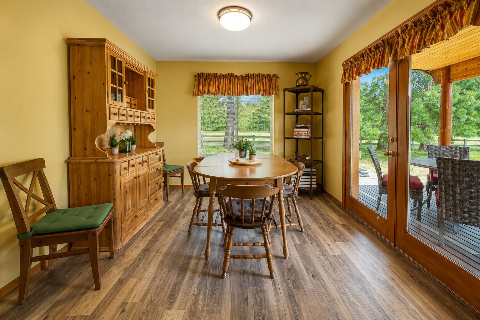 Un comedor con mesa y sillas de madera, un aparador con puertas de cristal, una estantería y vistas al exterior.