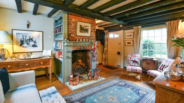 Uma sala de estar acolhedora com vigas de madeira aparentes, lareira de tijolos, estante de livros e uma grande janela com cortinas.