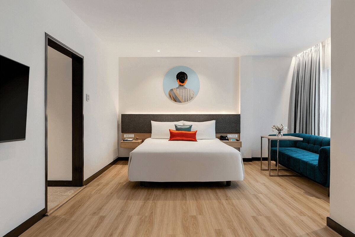 A modern hotel room with a large bed, a bedside table, a nightstand, a small table with a vase, and a blue sofa.