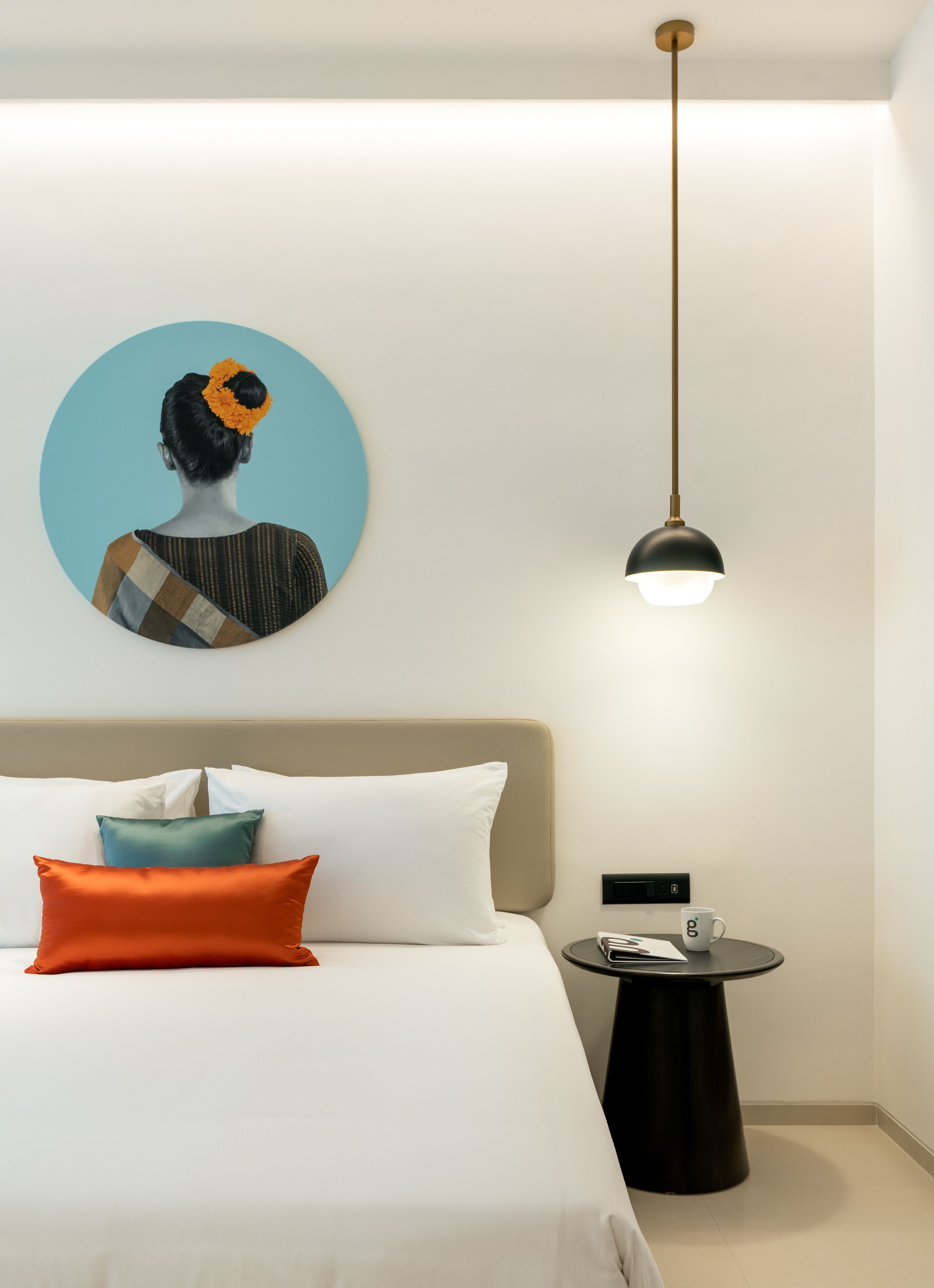 Una habitación de hotel moderna con una cama, almohadas, una mesa redonda con una taza y una lámpara colgante.