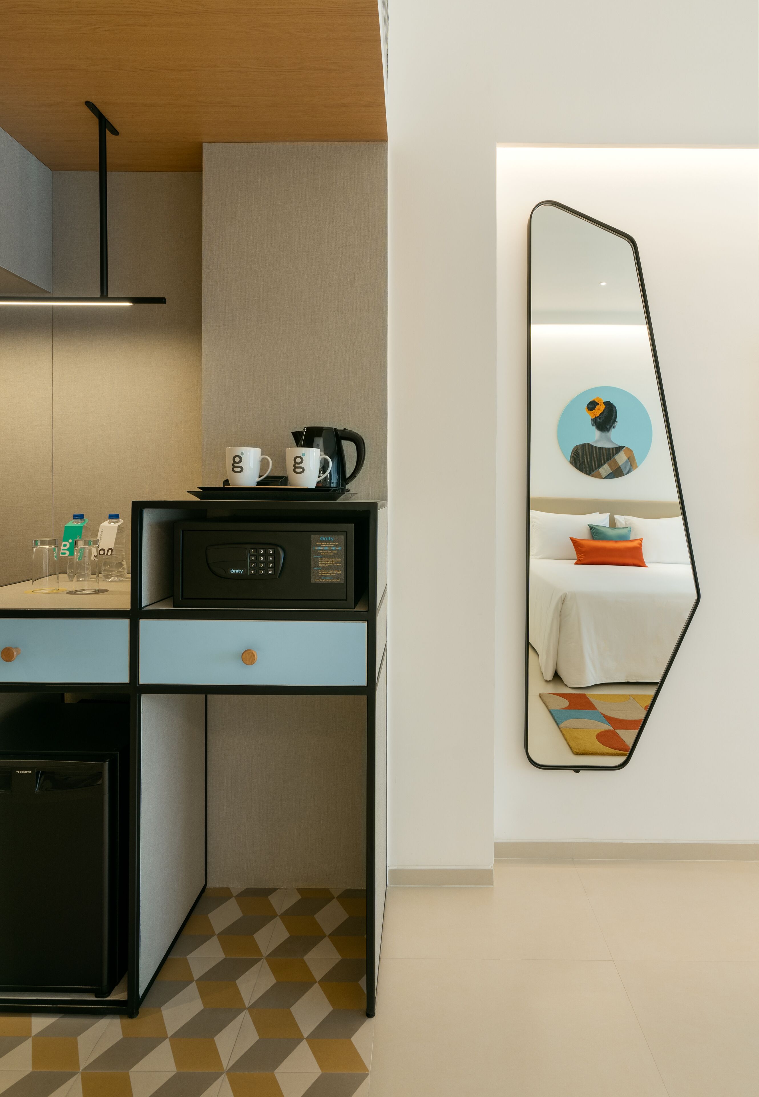 A compact hotel room with a kitchenette, a bed with a white duvet and orange pillow, a large mirror, and a wall-mounted artwork.