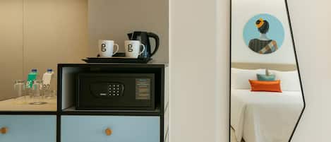 A compact hotel room with a kitchenette, a bed with a white duvet and orange pillow, a large mirror, and a wall-mounted artwork.