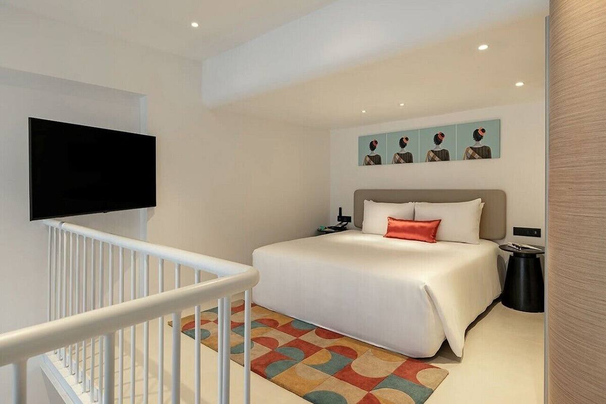 A modern hotel room with a large bed, a flat-screen TV mounted on the wall, a small bedside table, and a colorful rug.