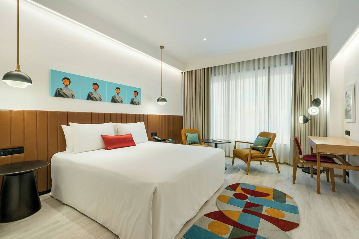 A hotel room with a large bed, a desk, a chair, a small table, and a wall-mounted TV.