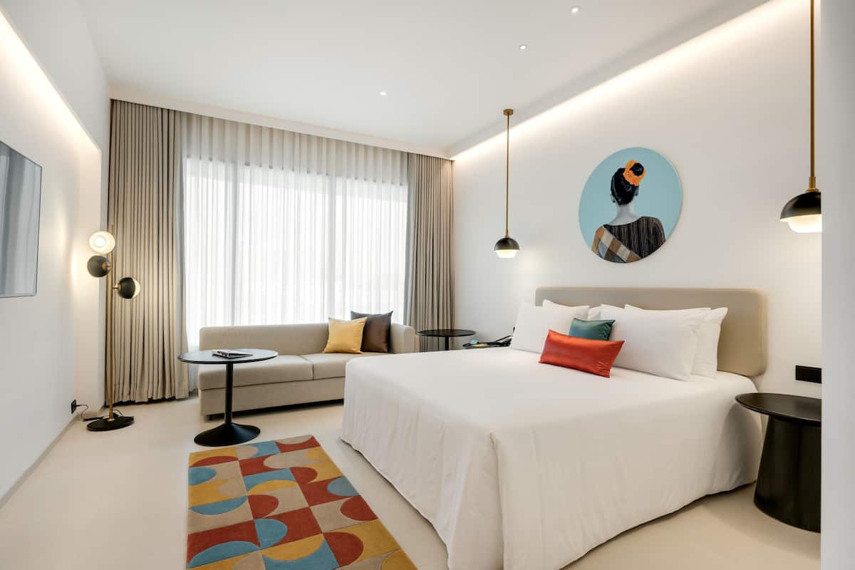 A modern hotel room with a large bed, a sofa, a small table, and a large window with curtains.