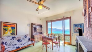 A living area with a sofa, dining table, chairs, a ceiling fan, and a view through a large window. - La Finestra Sul Lago: Lake Maggiore View & Garden (Stresa)