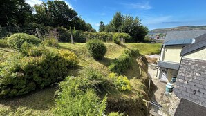 A hillside garden with various shrubs and ferns, a wooden fence, and a house with a sloped roof.