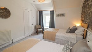 A bedroom with two beds, a chair, a radiator, a window with curtains, and a wall-mounted light fixture.
