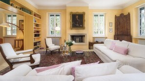 Un salon avec une cheminée, des canapés blancs, une bibliothèque et un tableau accroché au mur.