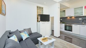 A modern living room with a grey sofa, a small white table, and a kitchen area with white cabinets and a black oven. - Apartment Denda (Dubrovnik)