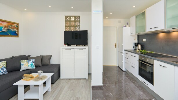 A modern apartment with a living area featuring a sofa, a coffee table, and a TV. The kitchen has white cabinets, a stove, and a countertop. - Apartment Denda (Dubrovnik)