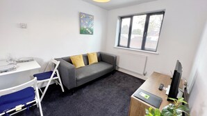 Een compacte, moderne kamer met een grijze bank, een witte tafel, een blauwe stoel, een radiator, een raam met jaloezieën en een kleine plant.