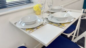 Mesa de comedor dispuesta con platos, fuentes y cubiertos blancos, junto con dos copas de vino y flores amarillas.