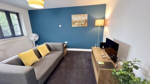 Una sala de estar moderna con un sofá gris, cojines amarillos, una pared azul decorativa, un cuadro, una mesita de madera, un televisor y una planta.