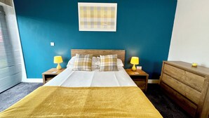 A hotel room with a bed, two bedside tables with lamps, a wooden dresser, and a blue wall with a framed picture. - 7 Cross Court, Stafford (Stafford)