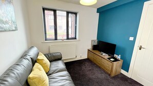 A living room with a leather sofa, a television on a wooden stand, a window with blinds, and a blue accent wall. - 7 Cross Court, Stafford (Stafford)