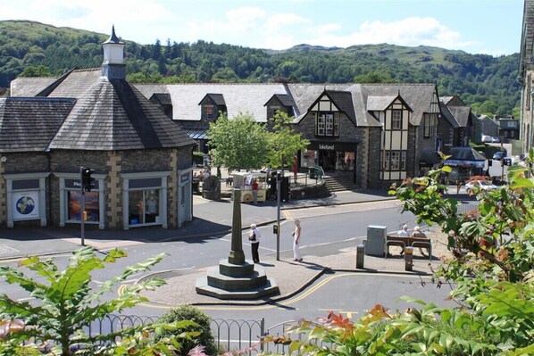 Une place de ville avec un monument central, des boutiques et, en arrière-plan, une vue sur des collines verdoyantes.