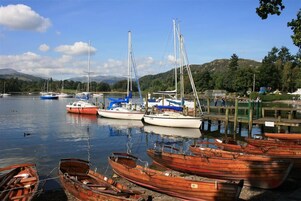 A marina with various boats docked, including sailboats and rowboats, surrounded by a calm lake and hills in the background.
