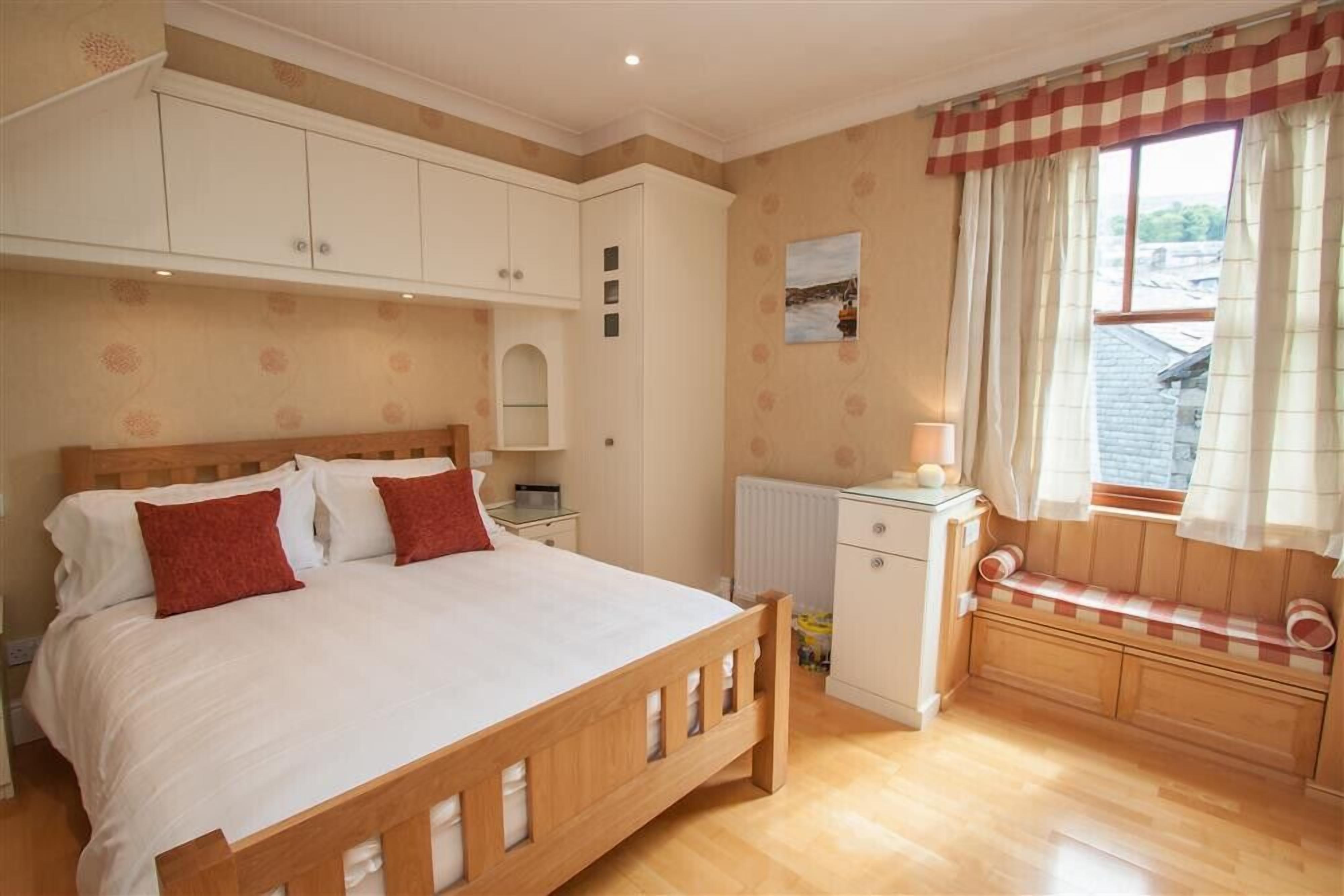 A bedroom with a wooden bed, white cabinets, a radiator, a bench by the window, and a small table with a lamp.