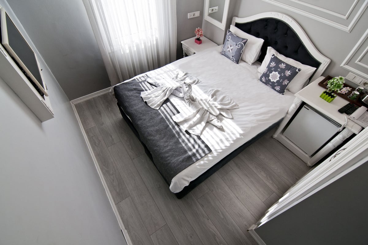 A neatly made bed with a dark headboard, a bedside table with a vase of flowers, and a small desk with a lamp and a plant.