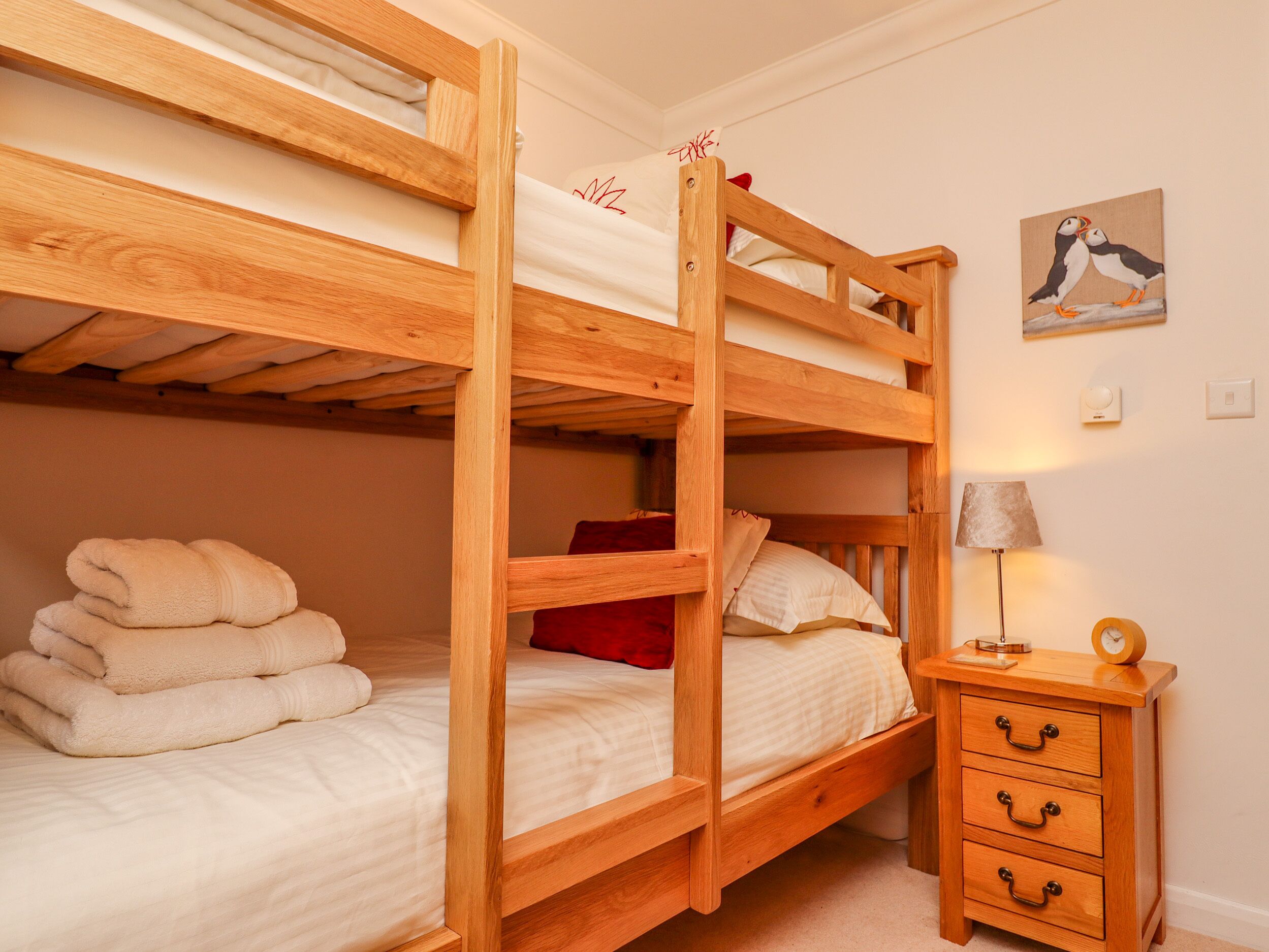A bunk bed with a wooden frame, a bedside table with drawers, a lamp, and a painting of birds on the wall.