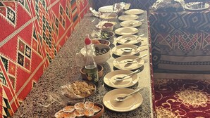 Uma mesa comprida com diversos alimentos e pratos, rodeada por tapetes estampados e uma toalha decorativa.