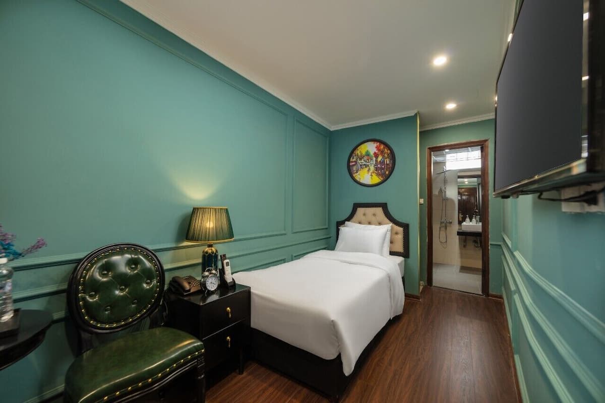 Een hotelkamer met turquoise wanden, een bed met witte lakens, een groene fauteuil, een nachtkastje met een lamp en een televisie.