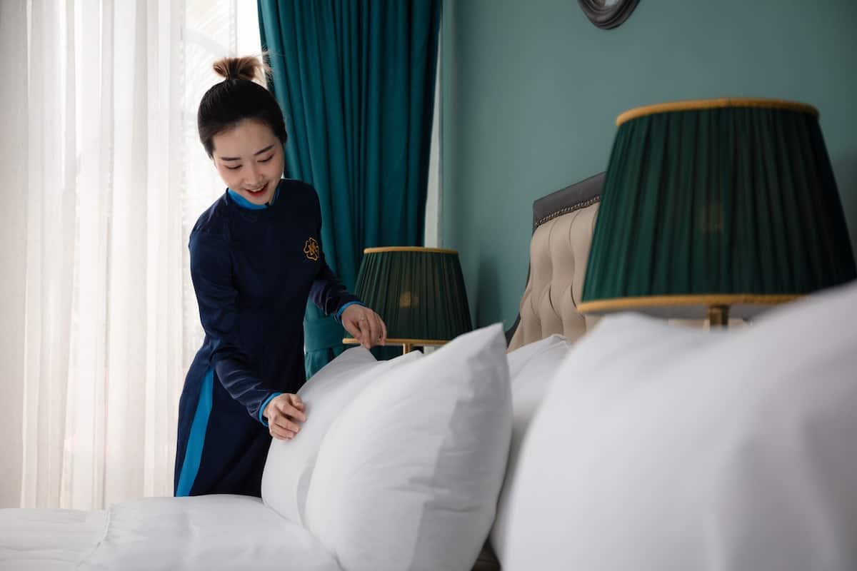 Een hotelkamer met een bed, een groene lamp en een persoon die de kussens opmaakt.