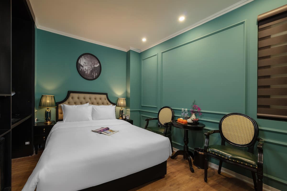 Een netjes ingerichte hotelkamer met een groot bed, een nachtkastje met een lamp, een stoel en een kleine tafel met een bloemenvaas.