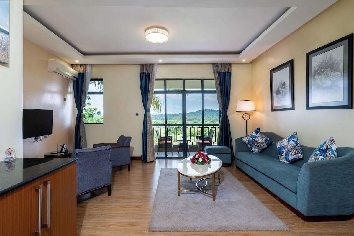 Ein modernes Wohnzimmer mit einer Couch, Sesseln, einem Couchtisch und einem Fernseher. Ein Balkon bietet Blick auf Grünflächen.