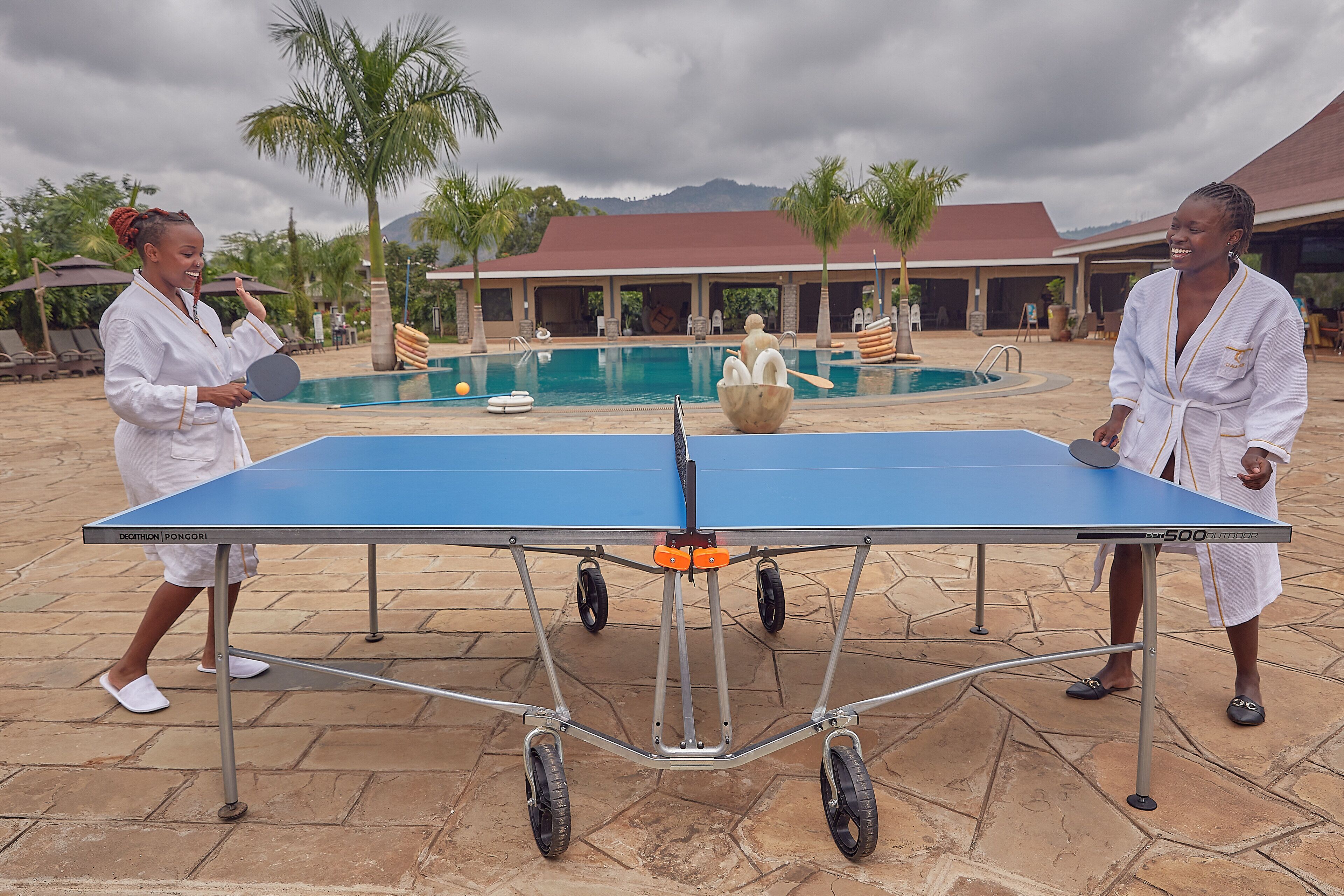 Two people playing table tennis outdoors near a pool with palm trees and a building in the background.