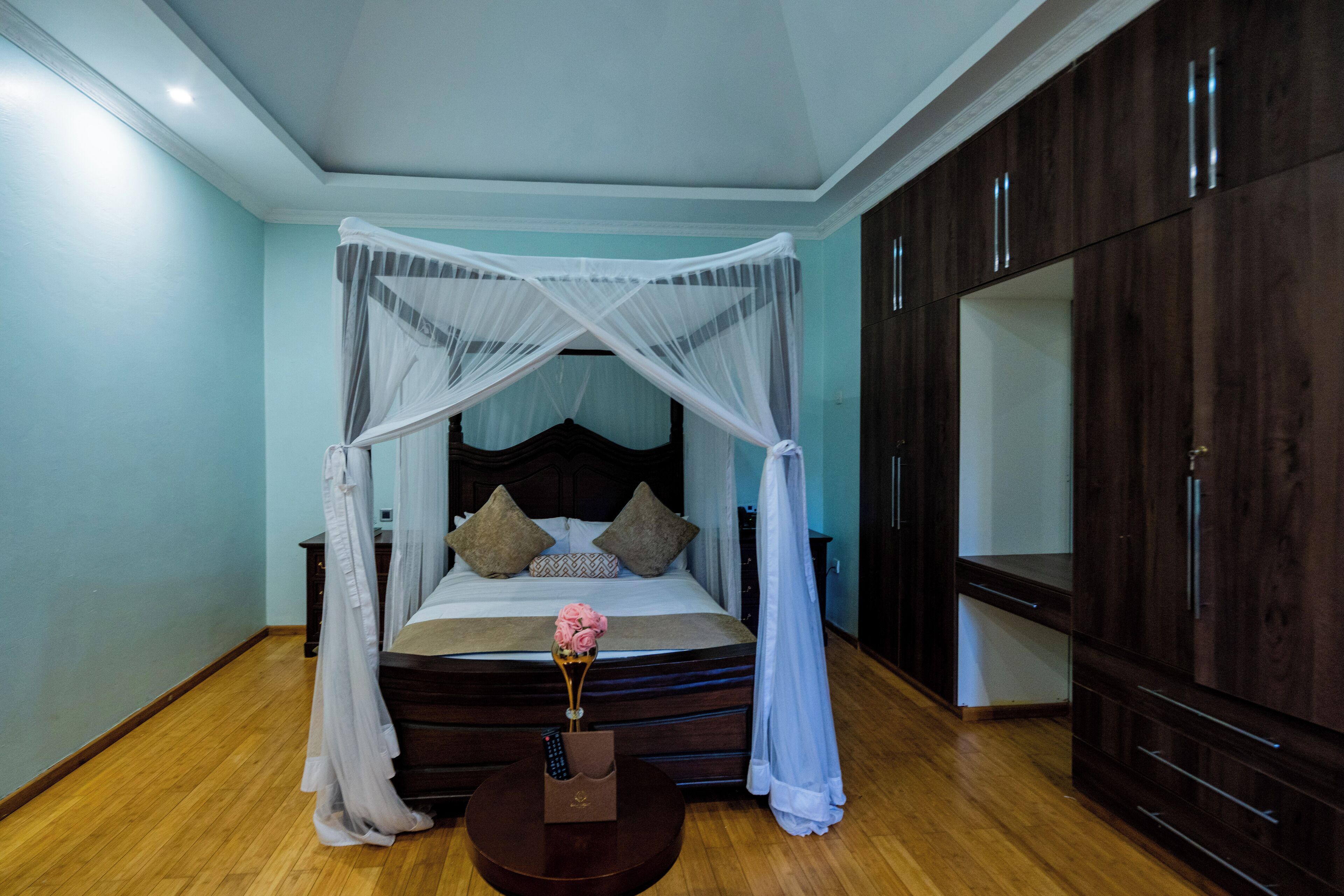 Un dormitorio con cama de dosel, suelo de madera, una mesita de noche con un florero y armarios de madera oscura.